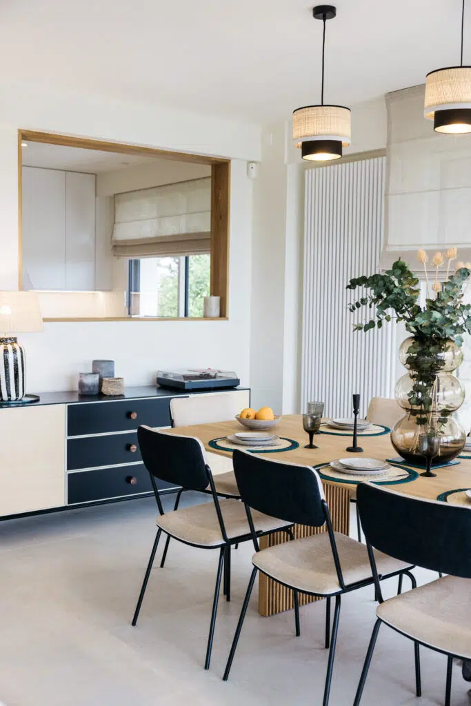 Coin repas avec table en bois, chaises à structure noire et assise claire, suspensions en tissu et buffet bas noir et blanc, ouvert sur la cuisine