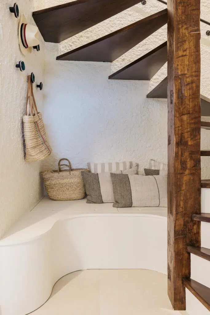 Banquette blanche aux formes arrondies sous un escalier en bois sombre, avec coussins en lin rayé, paniers en osier et patères murales
